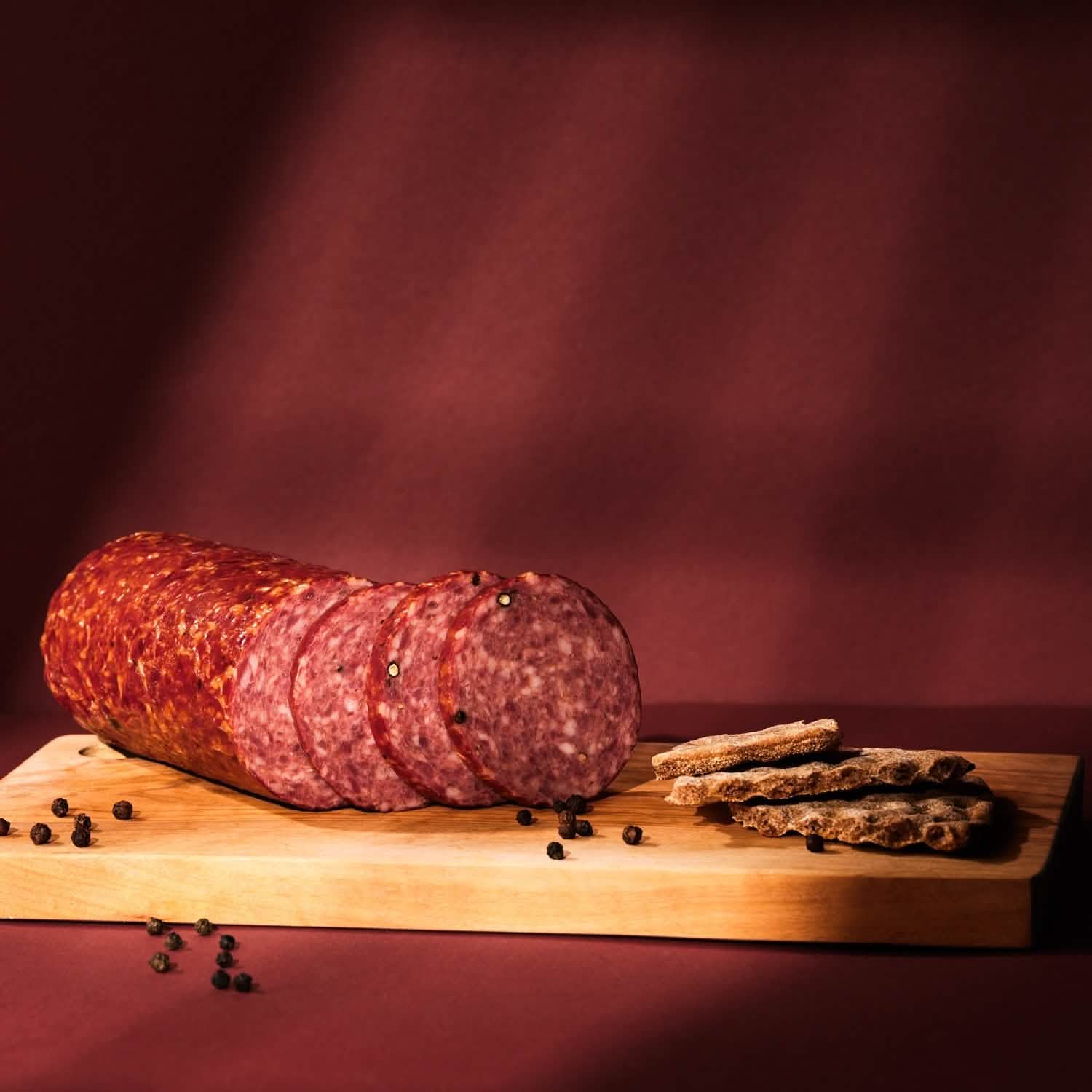 Salami-Strang angeschnitten mit Scheiben, Getreidecrackern und Pfefferkörner auf Holzbrett.
