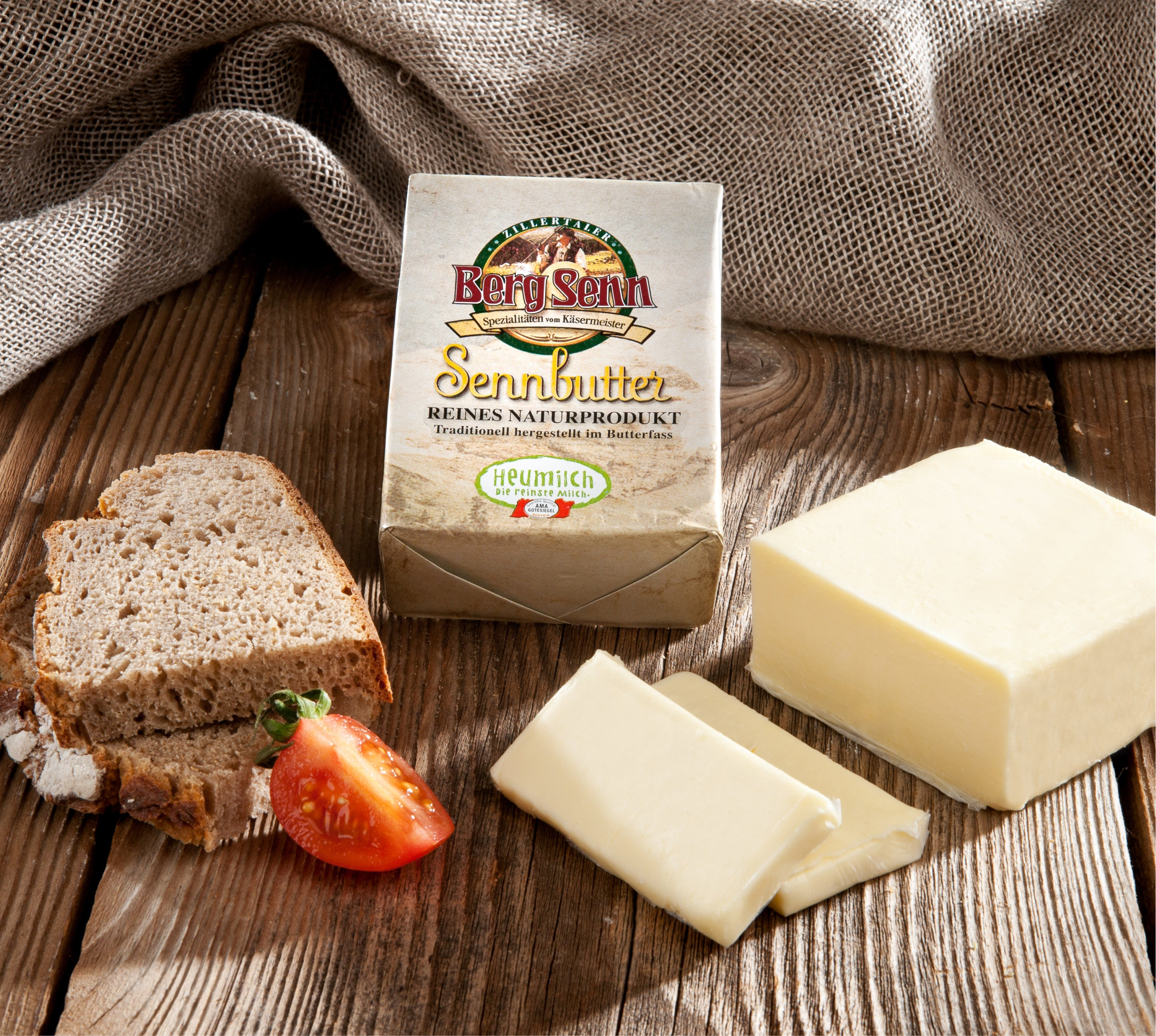 Butterstücke und Brotscheibe mit Berg Senn Butterverpackung auf Holzbrett und Leinenstoff.