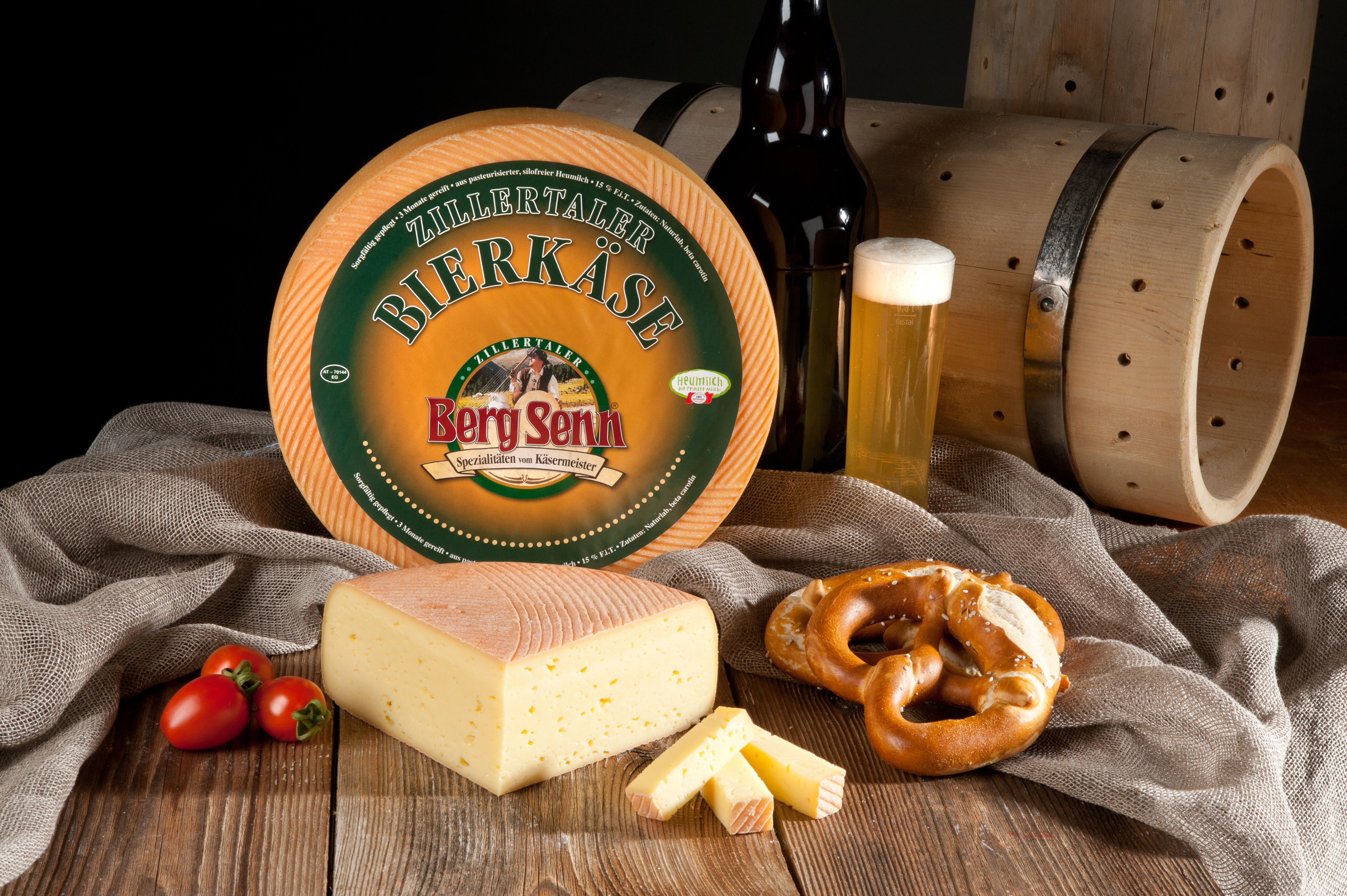 Bierkäse mit Anschnitt, Brezel, Bier und Holzfässer auf Holztisch mit Leinentuch.