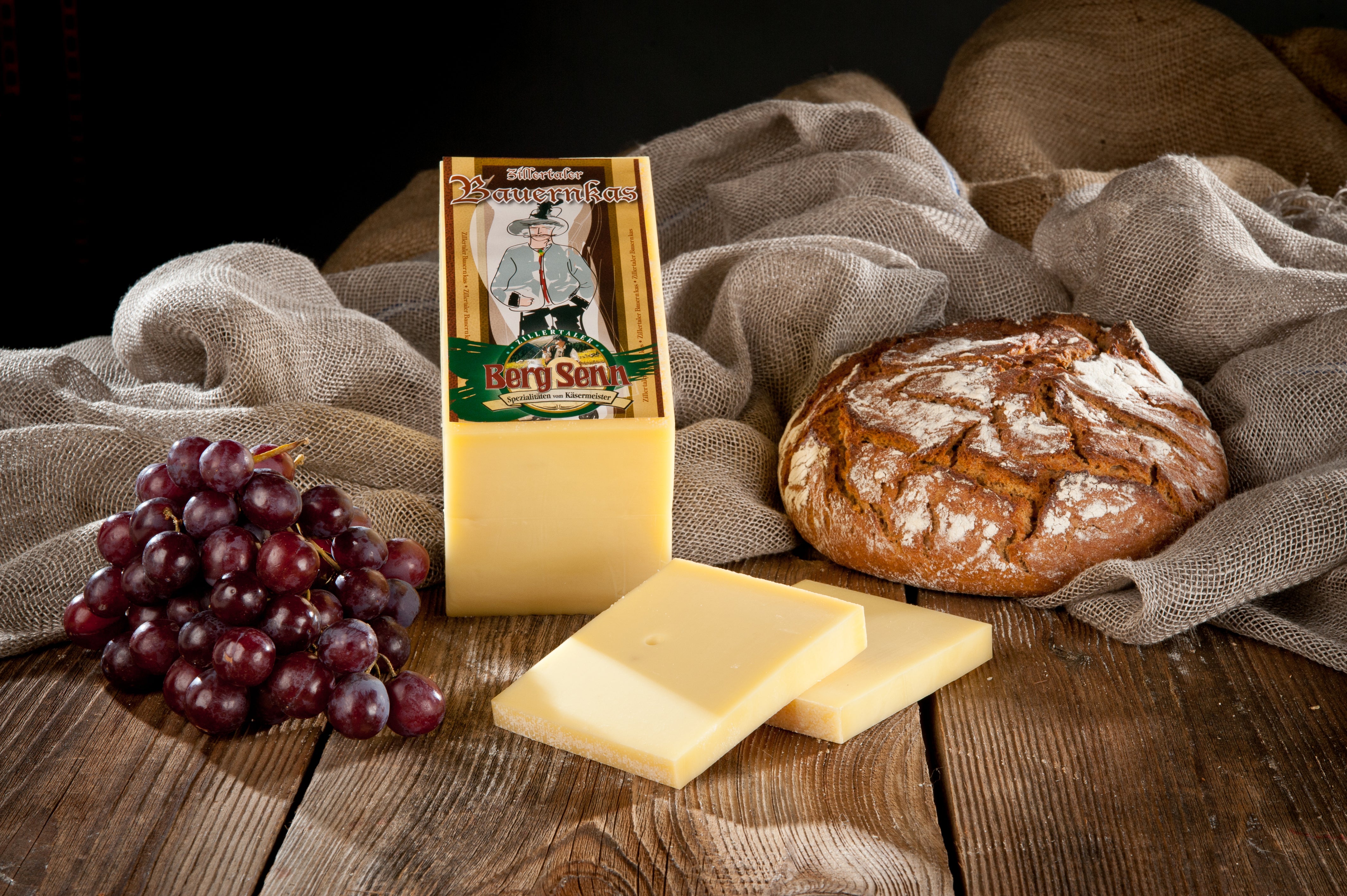 Käseplatte mit Bergkäse-Packung, Weintrauben, Brot und Leintüchern auf Holztisch.