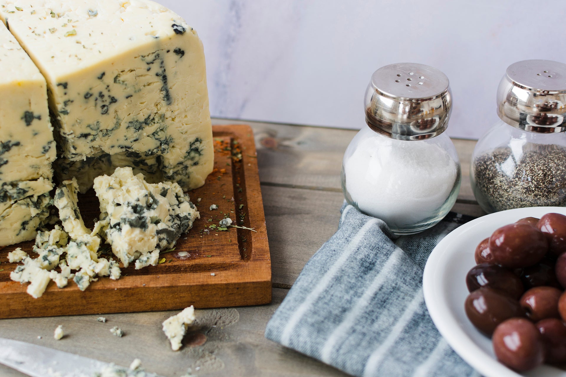 Blauschimmelkäse angeschnitten auf Holzbrett mit Salz, Pfeffer und roten Oliven.